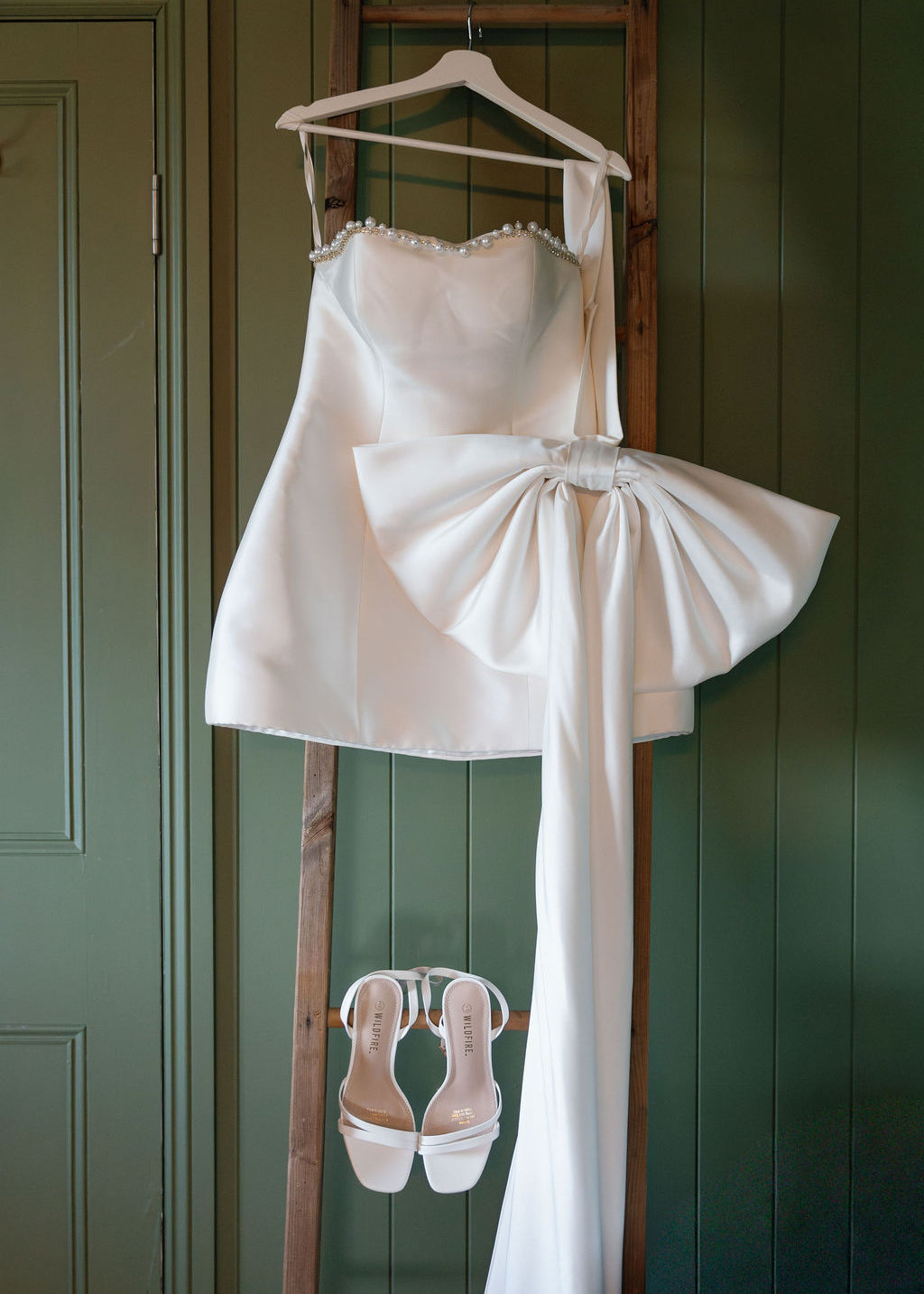 White wedding dress with a large bow and matching shoes hanging on a wooden rack against a green wall.