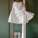 White wedding dress with a large bow and matching shoes hanging on a wooden rack against a green wall.