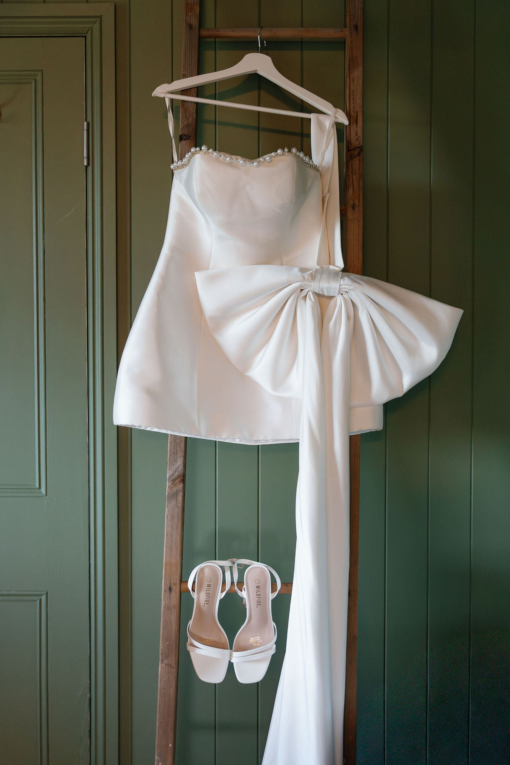 White wedding dress with a large bow and matching shoes hanging on a wooden rack against a green wall.